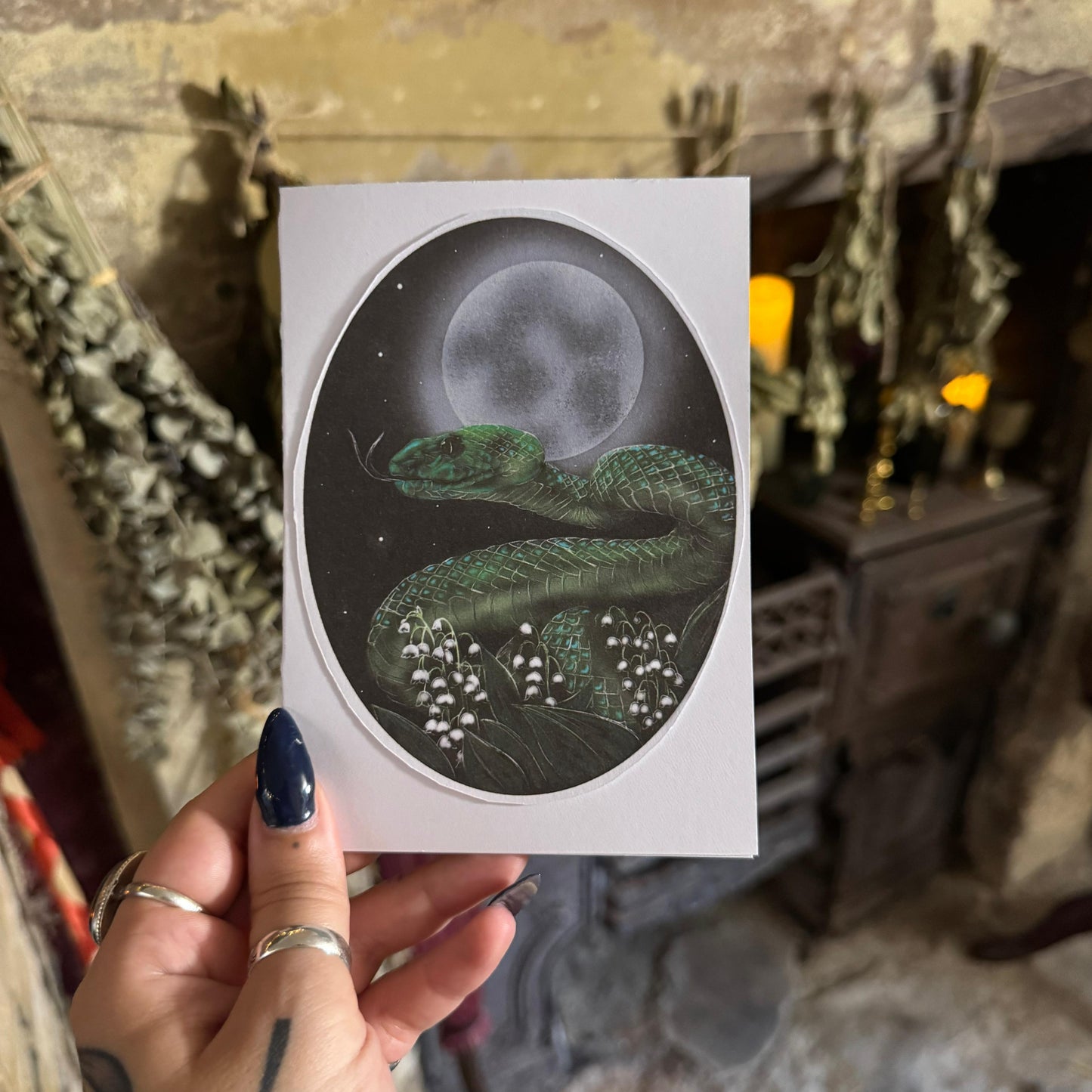 Hand holding a card with a snake and moon illustration against a rustic background.