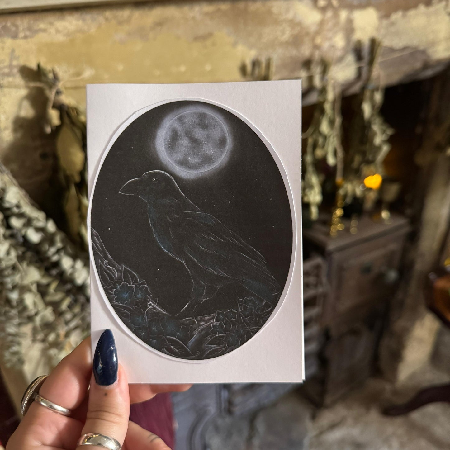 Sticker of a crow and moon design held by a hand with dark nail polish.
