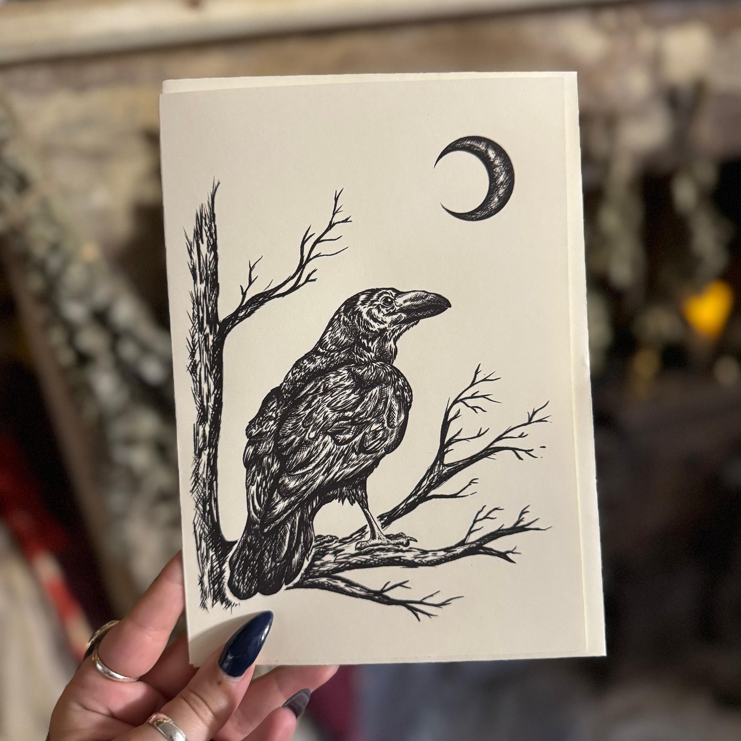 Card with a black and white illustration of a crow on a branch with a crescent moon above, held by a person.