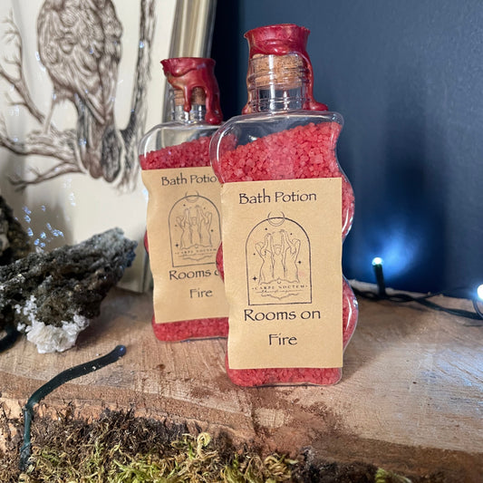 Bottle labeled 'Rooms on Fire' with a label featuring text and illustrations, placed on a wooden surface with moss.