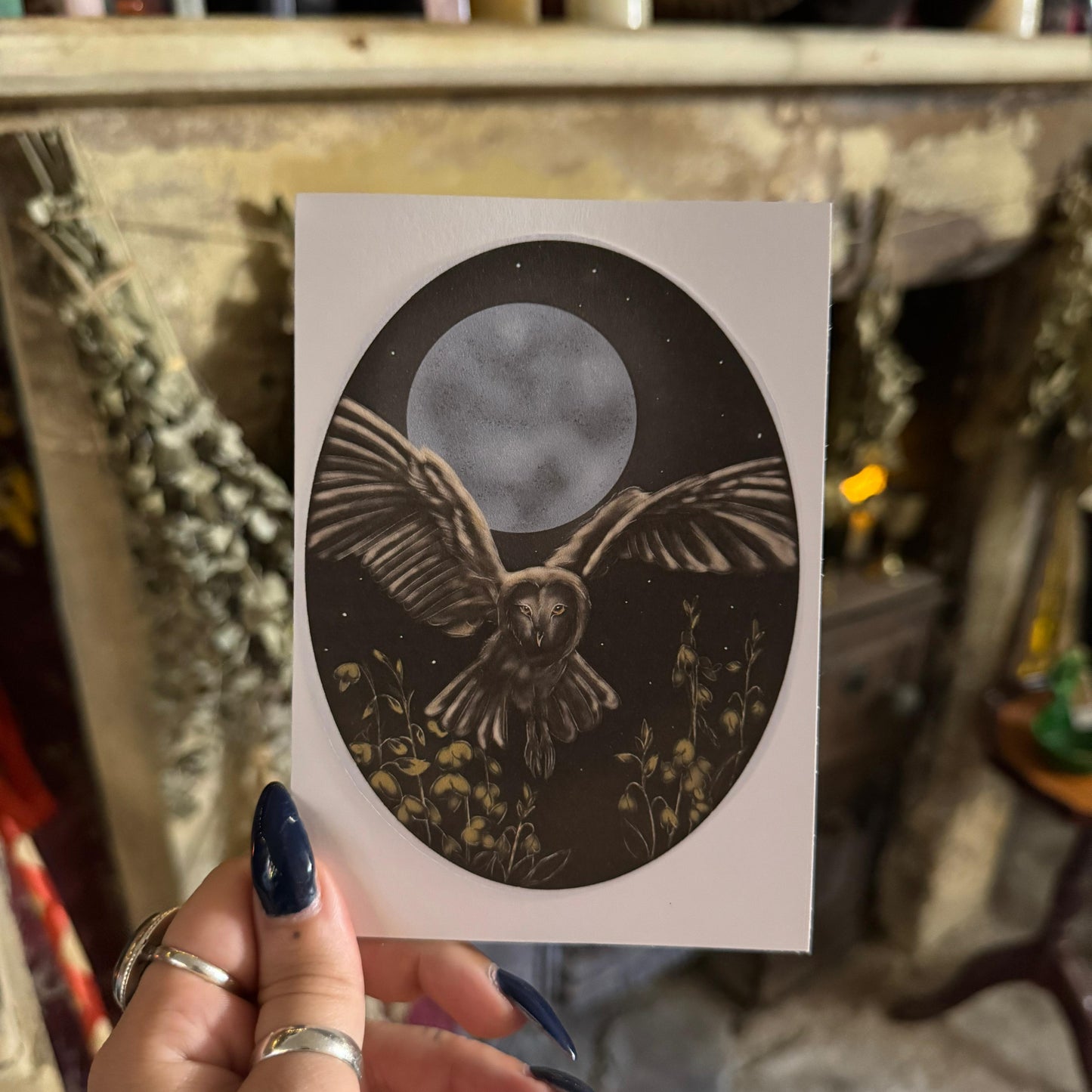 Hand holding a card with an owl design against a moonlit sky, with a blurred background of candles and decor.
