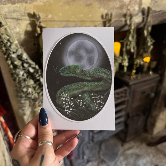 Hand holding a card with a snake and moon illustration against a rustic background.