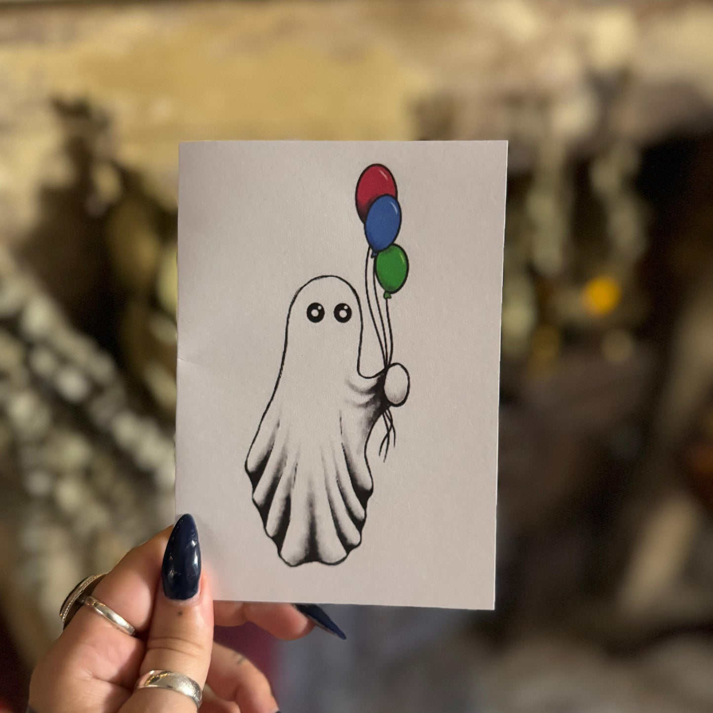 Hand holding a card with a black and white ghost holding balloons illustration.
