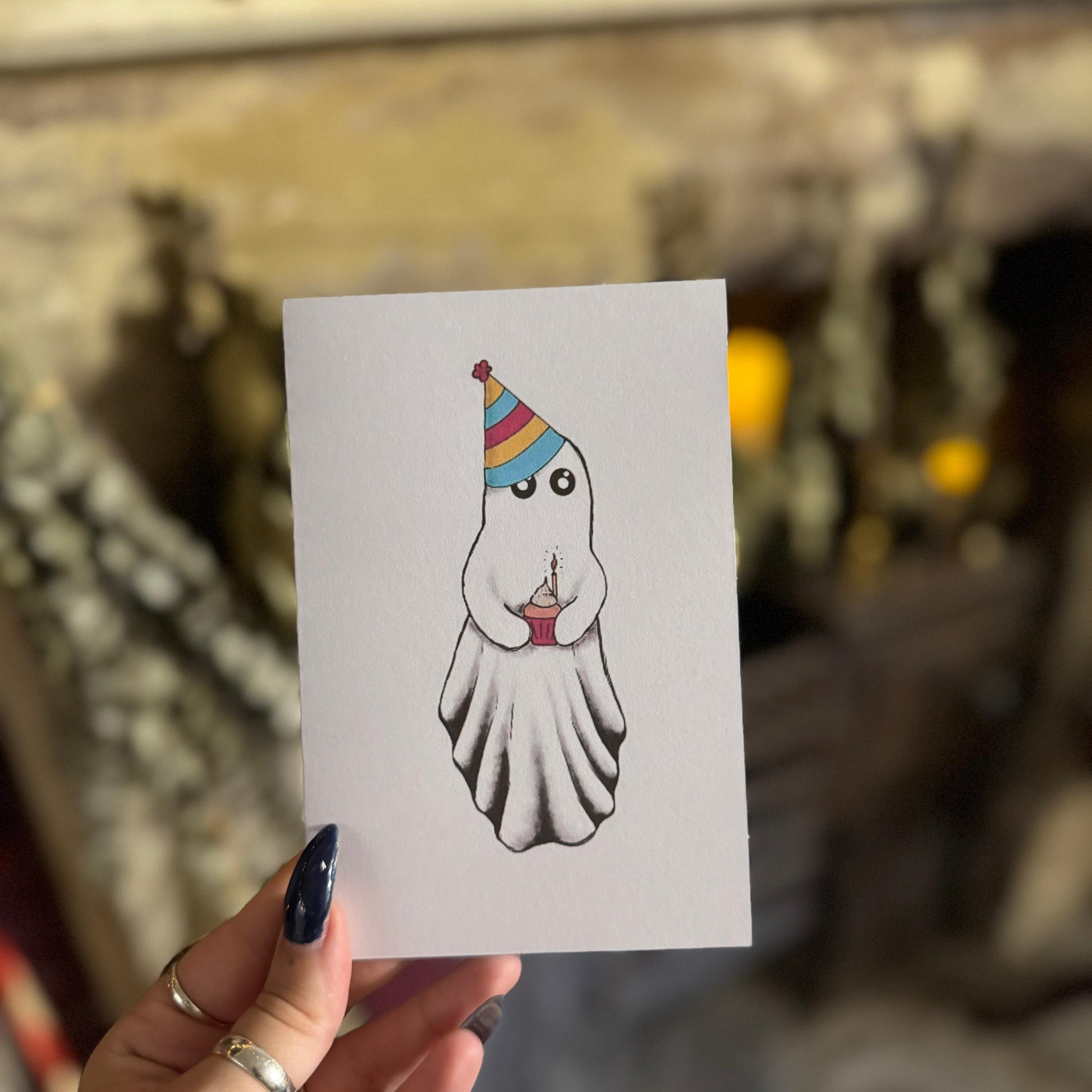 Hand holding a card with a cartoon ghost wearing a party hat against a blurred background