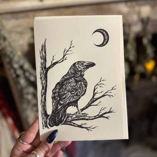 Card with a black and white illustration of a crow on a branch with a crescent moon above, held by a person.